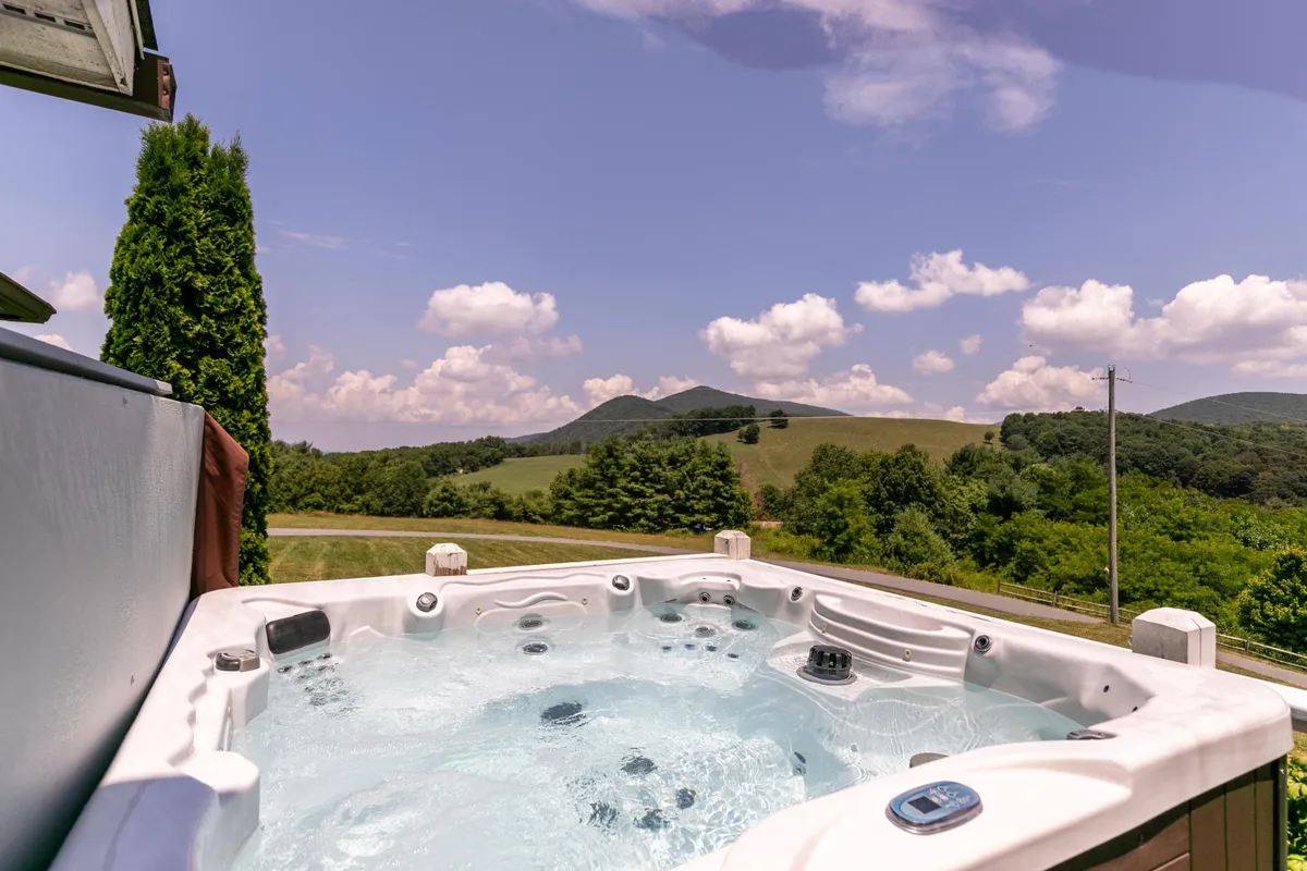 A serene outdoor hot tub set against a backdrop of lush greenery and distant mountains. The spacious deck provides room for relaxing, with ample seating and open sky views—perfect for soaking while watching the sun set or stargazing at night.