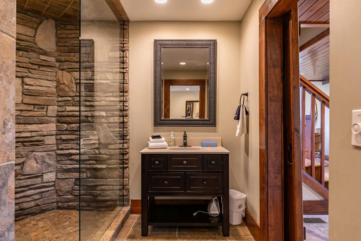 A serene bathroom with a glass shower enclosure, stone walls, and a dark wood vanity with a framed mirror. The natural materials evoke a rustic, spa-like feel that aligns with the glamping theme while highlighting modern comfort.