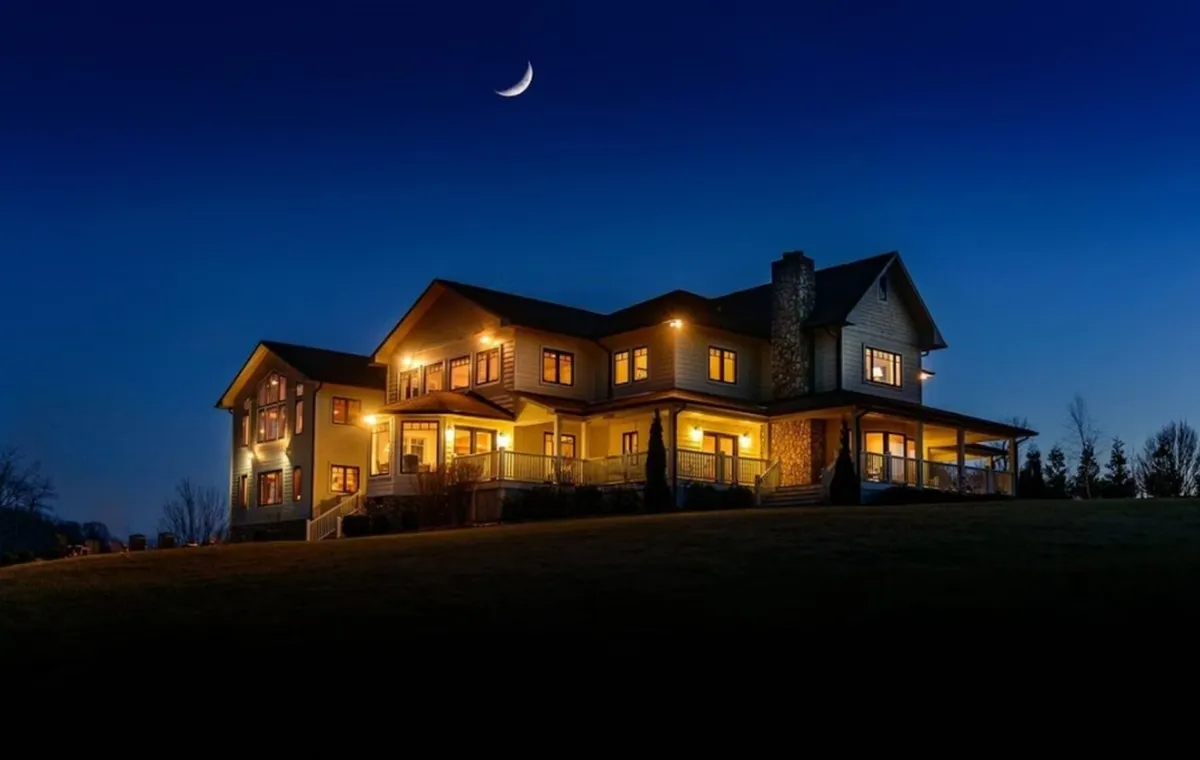 The image showcases the illuminated exterior of a spacious mountain home at dusk, with a striking stone chimney and a glowing, inviting porch. The serene Blue Ridge landscape surrounds the house, and a full moon rises in the evening sky, setting a tranquil, cozy ambiance ideal for gatherings and stargazing.