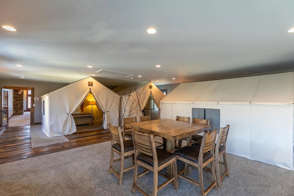 This image features a series of glamping-style canvas tents set up indoors, surrounded by a wooden dining table and chairs, evoking the feeling of an adventure camp within the comfort of home. The setting is bright and spacious, capturing the unique concept of indoor glamping while suggesting group activities and shared meals.