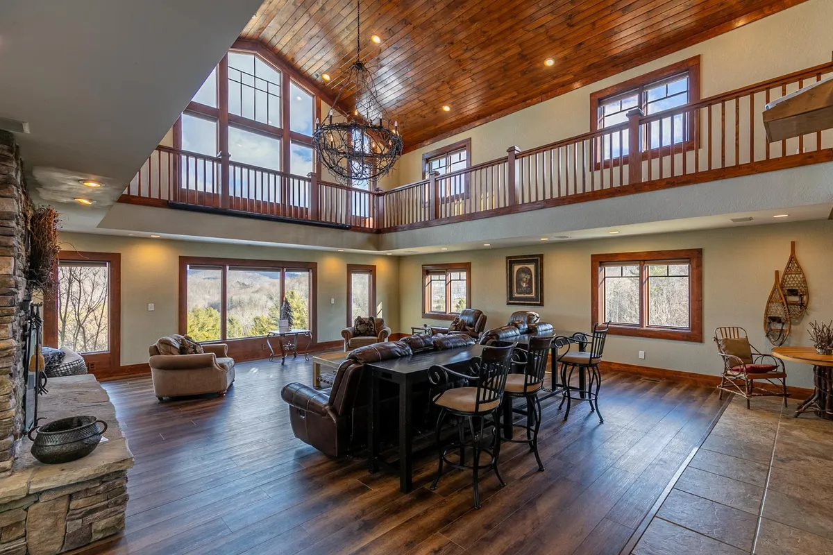 A cozy yet luxurious living area with a grand stone fireplace, a soaring wooden ceiling, a chandelier, generous seating, and large windows framing mountain vistas. The open floor plan makes the space feel connected and perfect for mingling.