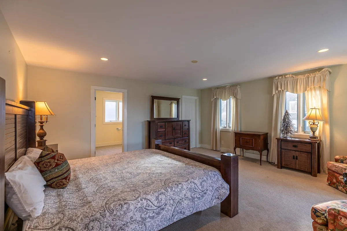 A tranquil bedroom with patterned bedding, a wooden dresser, an inviting armchair by the window, and soft natural light streaming in. The window hints at potential mountain views, contributing to a peaceful atmosphere.