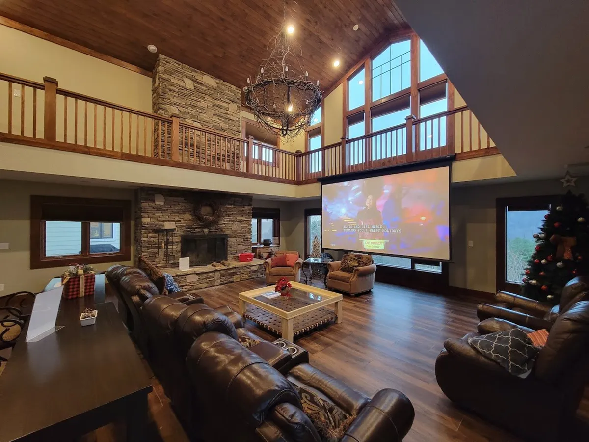 A living room arranged for movie nights, featuring a large roll-down projection screen, comfortable seating, a stone fireplace, and a warm, ambient chandelier. The room exudes comfort and togetherness, ideal for cozy evenings with family and friends.
