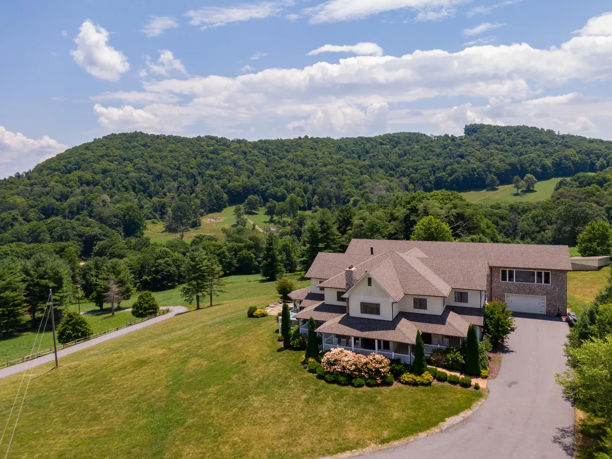 This image showcases the large farmhouse set amidst rolling hills and lush greenery, with a clear view of the driveway leading up to the property. The setting sun casts a warm glow over the landscape, highlighting the tranquil, open space ideal for outdoor gatherings and nature walks.