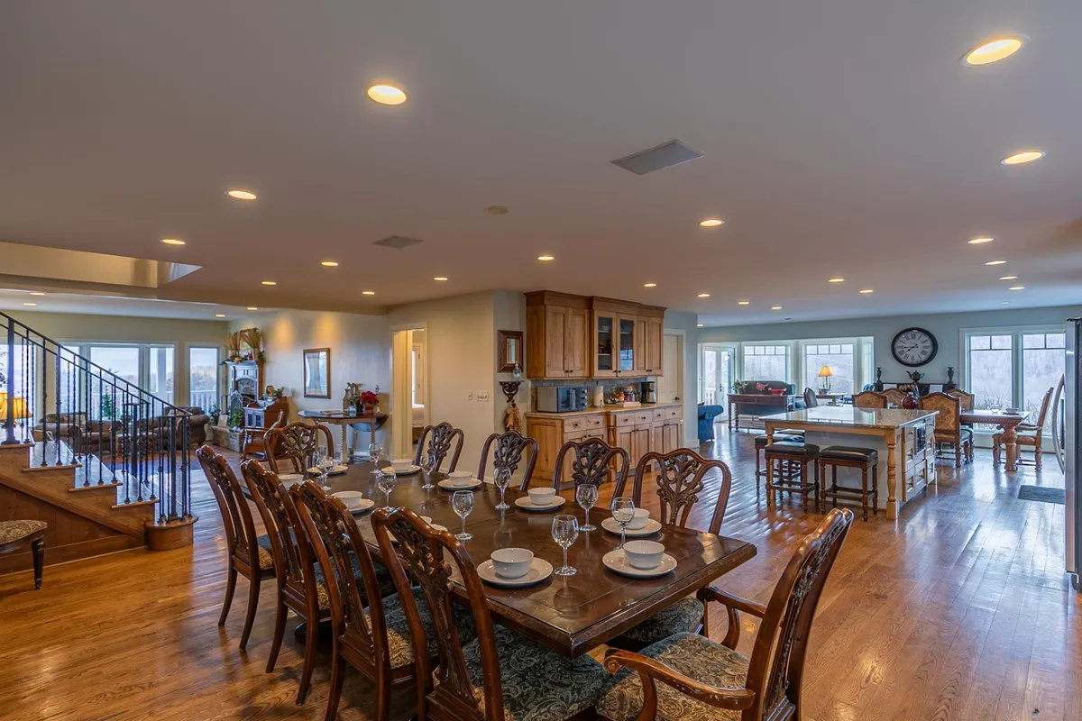 This image features a large dining table set for a gathering, an open kitchen visible in the background, a staircase leading to an upper level, and expansive windows showcasing scenic views. The space feels warm, inviting, and well-suited for hosting large groups.